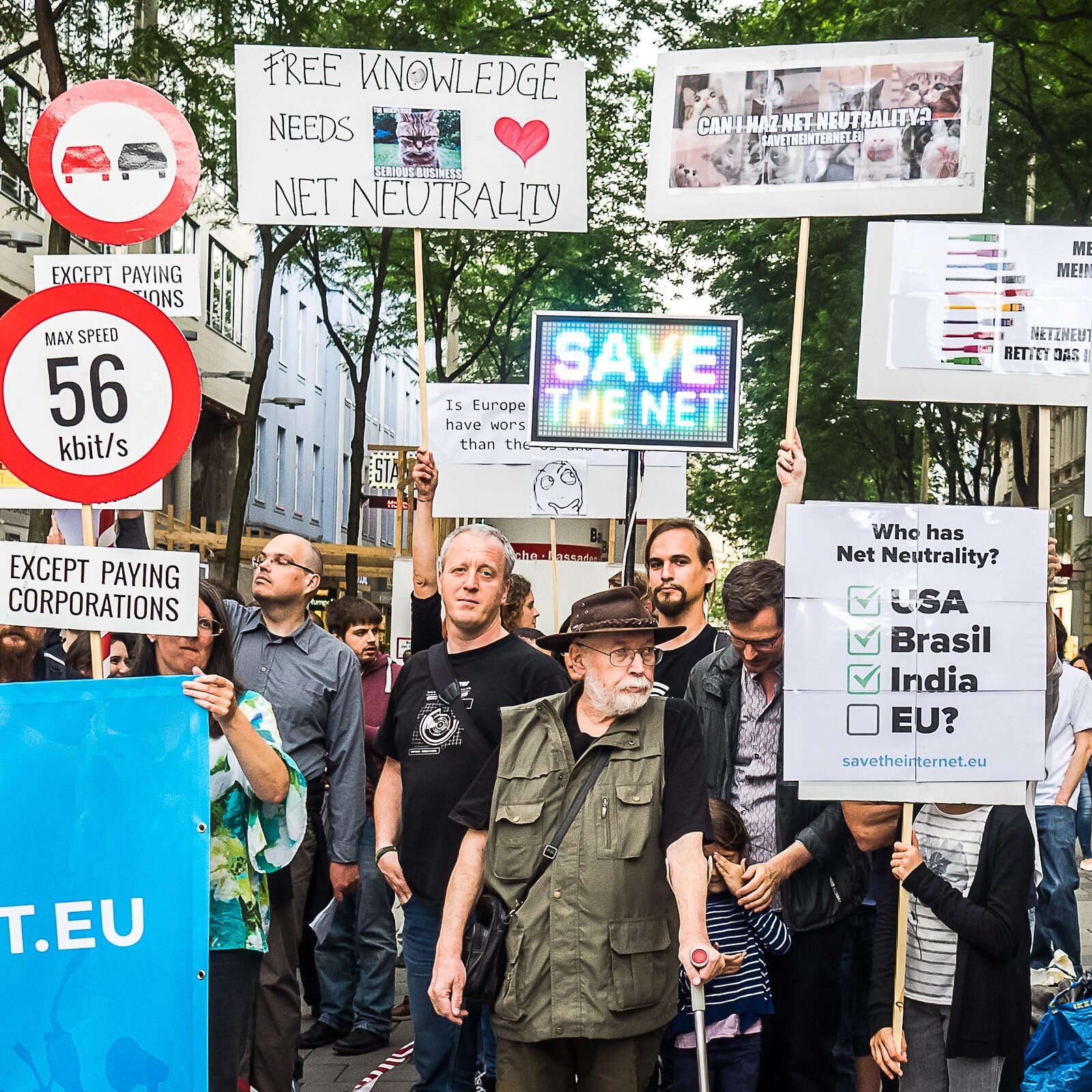 Crowd at the 2016 Vienna save-the-internet protest holding signs including “Free knowledge needs net neutrality” and a “56 kbit/s” speed-limit sign
